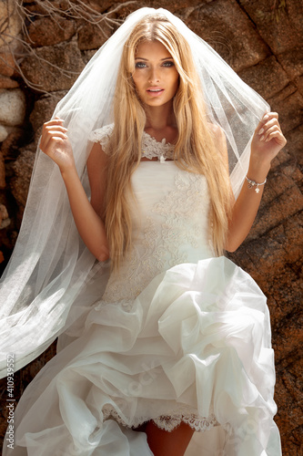 Beautiful sexy blonde woman in wedding dress with white veil and long hair and tanned body posing at the stone.