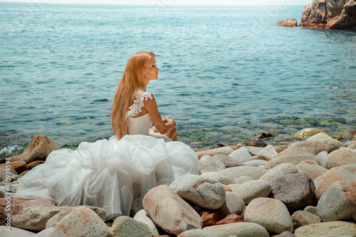 Obraz na płótnie Beautiful sexy blonde woman in wedding dress with long hair and tanned body sitting at the seashore. w salonie