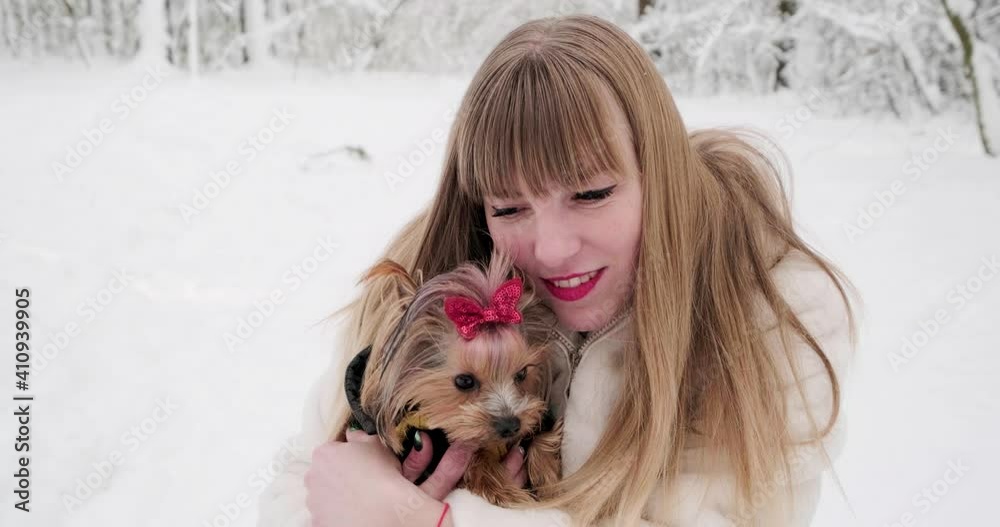 Beautiful smiling young woman in white fur coat looking at camera posing at snowy forest with yorkshire terrier small dog with red bow Beautiful smiling young woman in white fur coat looking at camera posing at snowy forest with yorkshire terrier small dog with red bow