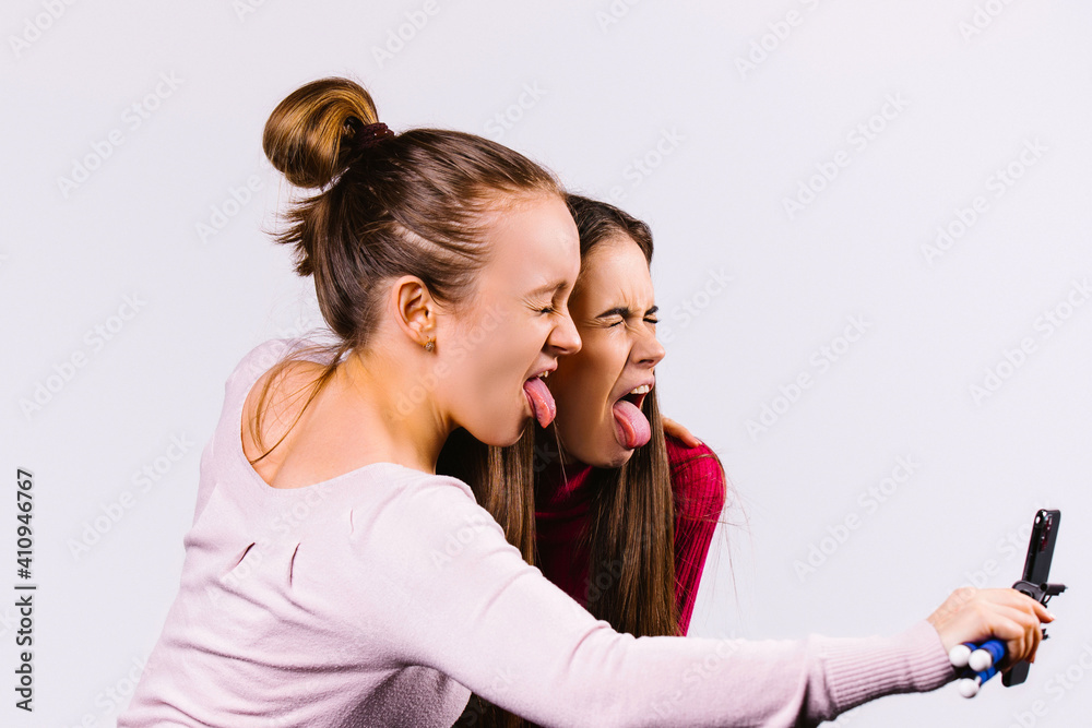 Two girlfriends fool around in live streaming with a smartphone on selfie stick show their tongues. Profile photo on white background. Emotion and pandemic pastime concept. Two girlfriends fool around in live streaming with a smartphone on selfie stick show their tongues. Profile photo on white background. Emotion and pandemic pastime concept.