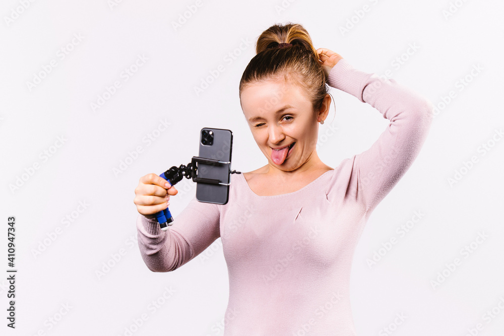 The blonde girl winks and shows her tongue while making a selfie on her smartphone. White background and playful mood. The blonde girl winks and shows her tongue while making a selfie on her smartphone. White background and playful mood.