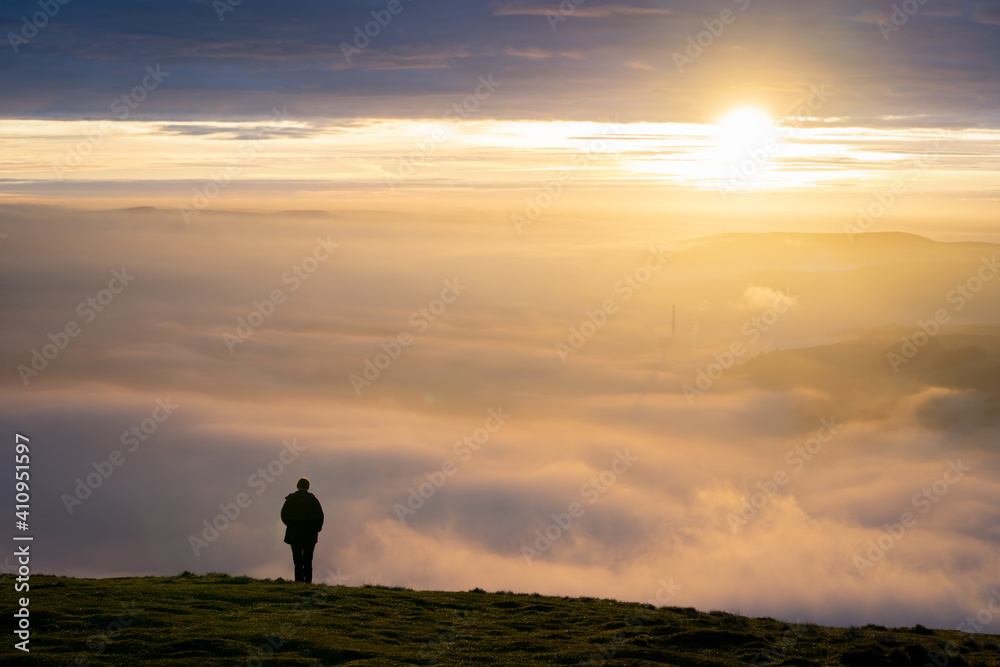 Silhouette of one lone man stood on edge of mountain peak top above clouds as fog mist and cloud inversion gathers watching the sun rise with a beautiful orange sunlight sky 