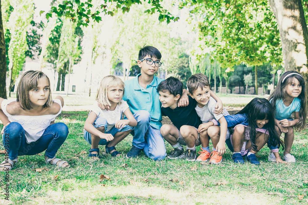 Fototapeta premium Row of happy kids doing squats together in park, hugging each other, looking away in excitement. Kids party or entertainment concept
