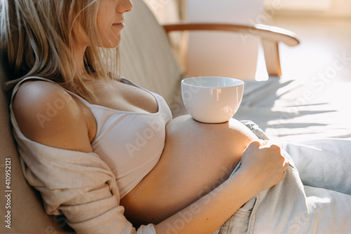 Pregnant woman sitting on a sofa with a white cup of coffee or tea on her belly, closeup.