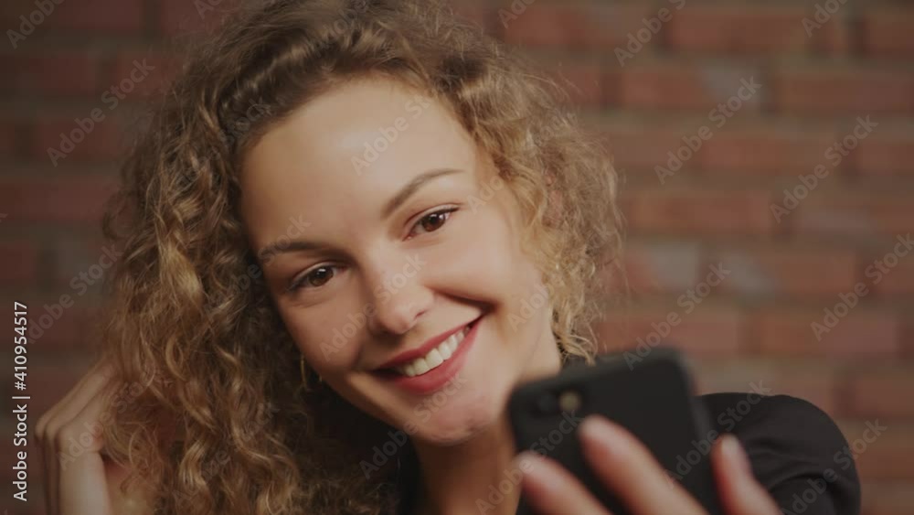 Portrait of a young curly caucasian business woman taking selfies and smiling with a red brick wall in a loft apartment Portrait of a young curly caucasian business woman taking selfies and smiling with a red brick wall in a loft apartment