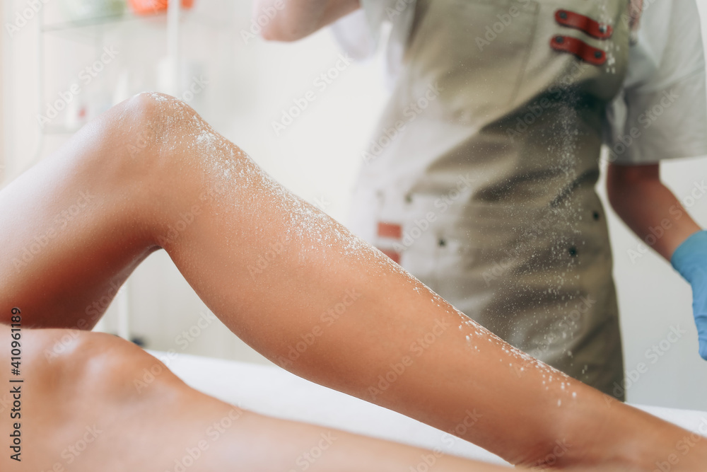 Fototapeta premium Beautician in an apron is sprinkling talcum powder on a young girl's leg before the epilation procedure. The girl is lying on a couch in a beauty salon, she does the procedure shugaring or waxing