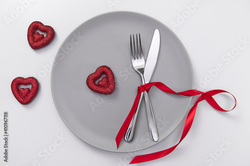 Valentine's day festive table setting, flat lay with red heart shape on gray plate, fork, knife and red ribbons on white table. Love dating concept. Place your text and copy space. High quality photo