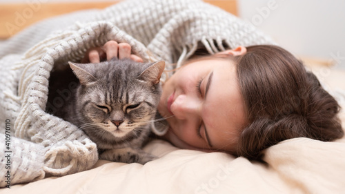 A teenage girl and a British cat lie under a blanket on the bed and sleep. Keeping warm in the cold winter Pets and friendship concept. Quarantine. Holidays. Stay home. Self-isolation. Social distance