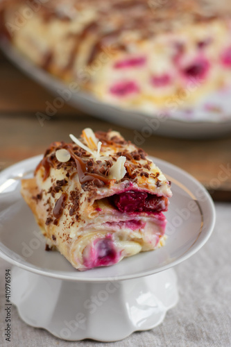 Cake from rolled pancakes with custard, cherries and grated chocolate on white plate, on a light background. Selective focus.