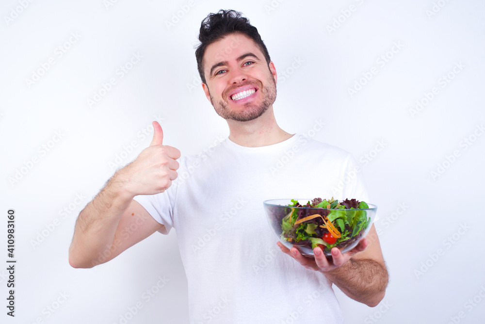 Young handsome Caucasian man holding a salad bowl against white background Waiving saying hello happy and smiling, friendly welcome gesture.