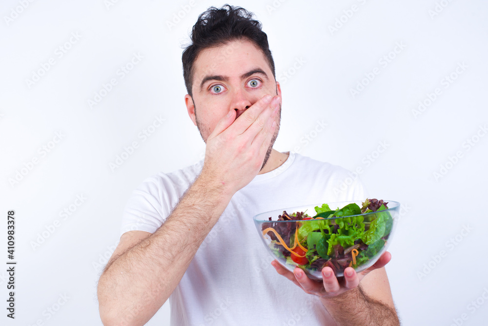 Young handsome Caucasian man holding a salad bowl against white background shocked covering mouth with hands for mistake. Secret concept.