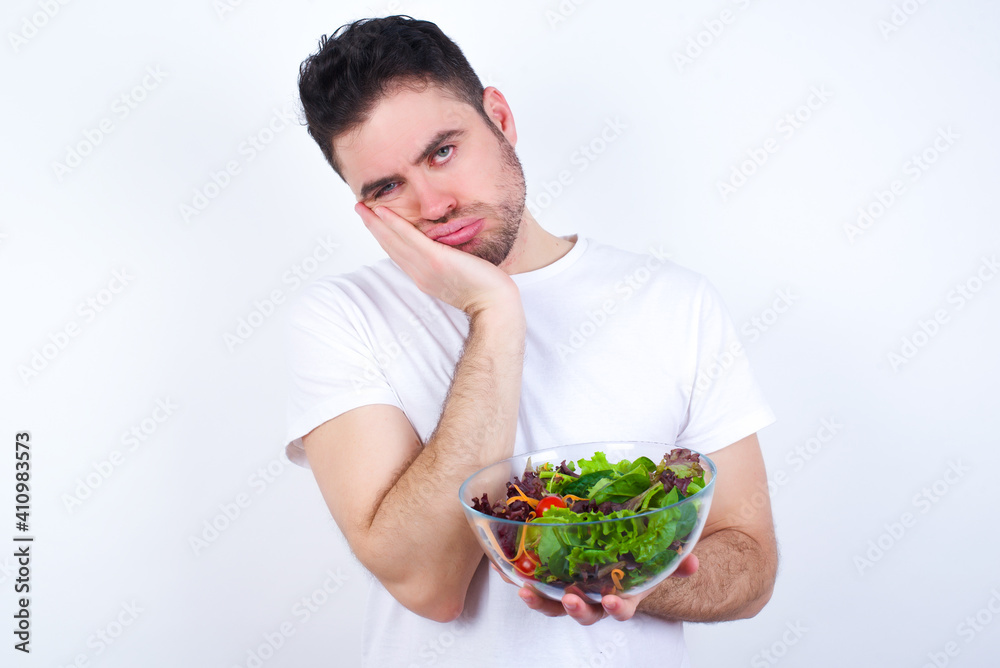 Very bored Young handsome Caucasian man holding a salad bowl against white background holding hand on cheek while support it with another crossed hand, looking tired and sick, Very bored Young handsome Caucasian man holding a salad bowl against white background holding hand on cheek while support it with another crossed hand, looking tired and sick,