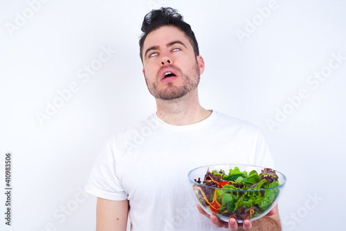 Young handsome Caucasian man holding a salad bowl against white background looking sleepy and tired, exhausted for fatigue and hangover, lazy eyes in the morning.