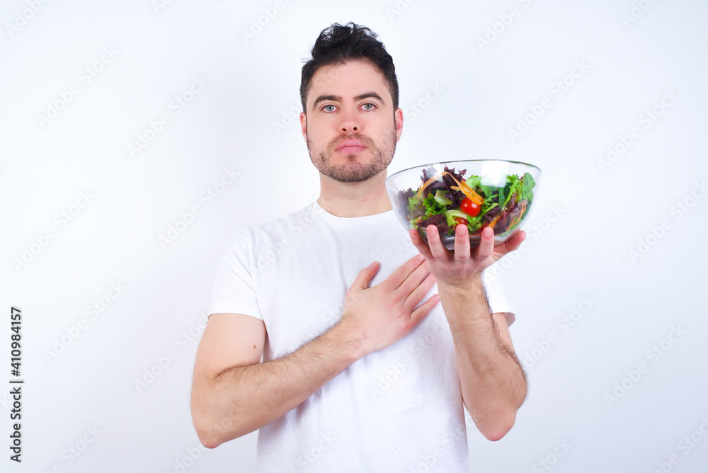 Young handsome Caucasian man holding a salad bowl against white background Swearing with hand on chest and open palm, making a loyalty promise oath Young handsome Caucasian man holding a salad bowl against white background Swearing with hand on chest and open palm, making a loyalty promise oath