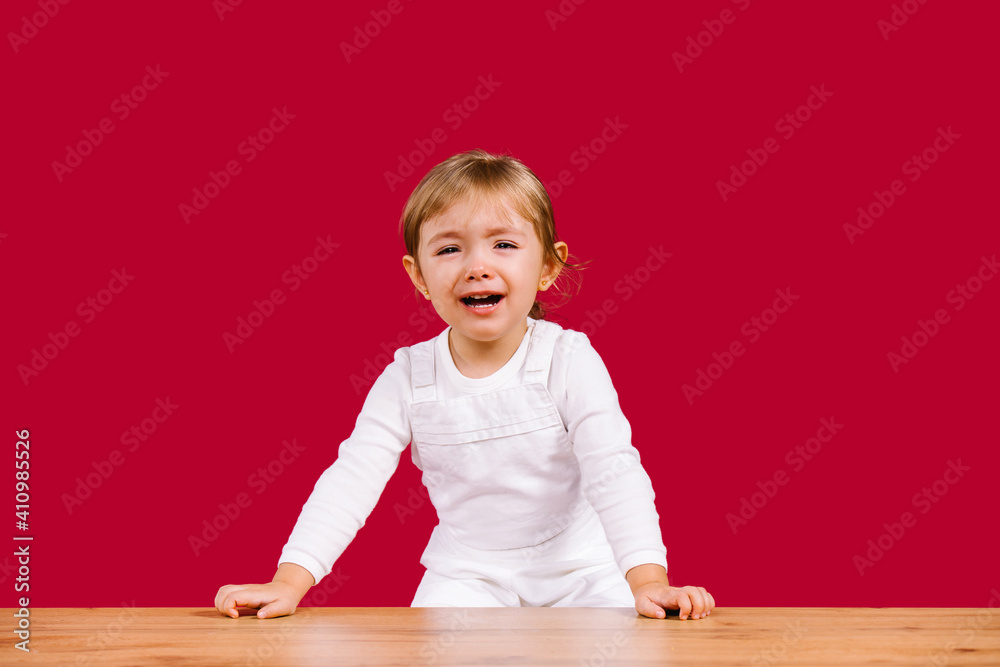 Naklejka premium Blonde toddler girl wearing casual white clothes is scared and crying girl standing over wooden table on red background