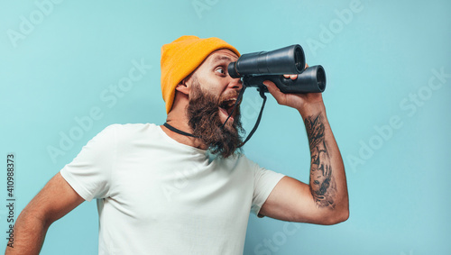 Young bearded hipster guy with binoculars over isolated blue wall wearing white blank t-shirt. Crazy emotions