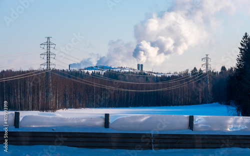 An electricity line with tall pylons standing on a snowy field. Two chimneys in the background.