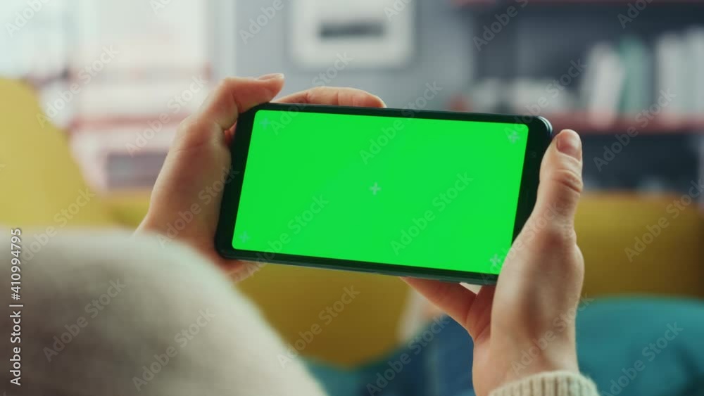 Beautiful Authentic Female Using a Smartphone with Green Screen Mock Up Display at Home Living Room while Lying on a Couch Sofa. She's Browsing the Internet and Checking Videos on Social Networks. Beautiful Authentic Female Using a Smartphone with Green Screen Mock Up Display at Home Living Room while Lying on a Couch Sofa. She's Browsing the Internet and Checking Videos on Social Networks.