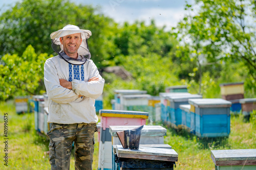 Beekeeper stands crosshands on the colorful hives background. Man wears protective hat with net and national shirt. Sunny day at the apiary.