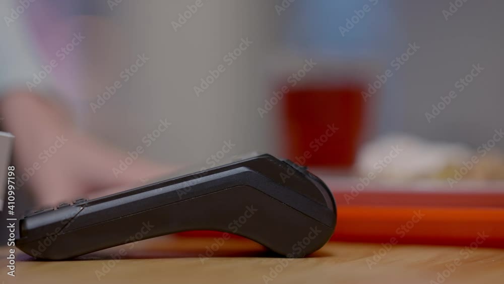 Side view of slim female hand paying with card on POS-terminal. Young Caucasian unrecognizable woman using cashless wireless payment in self-service restaurant and leaving with blurred food tray. Side view of slim female hand paying with card on POS-terminal. Young Caucasian unrecognizable woman using cashless wireless payment in self-service restaurant and leaving with blurred food tray.