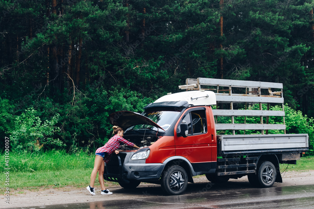 A young slender beautiful mechanic girl in a red checked shirt and short denim shorts repairs a gazelle car on the highway near the forest. Beautiful summer landscape