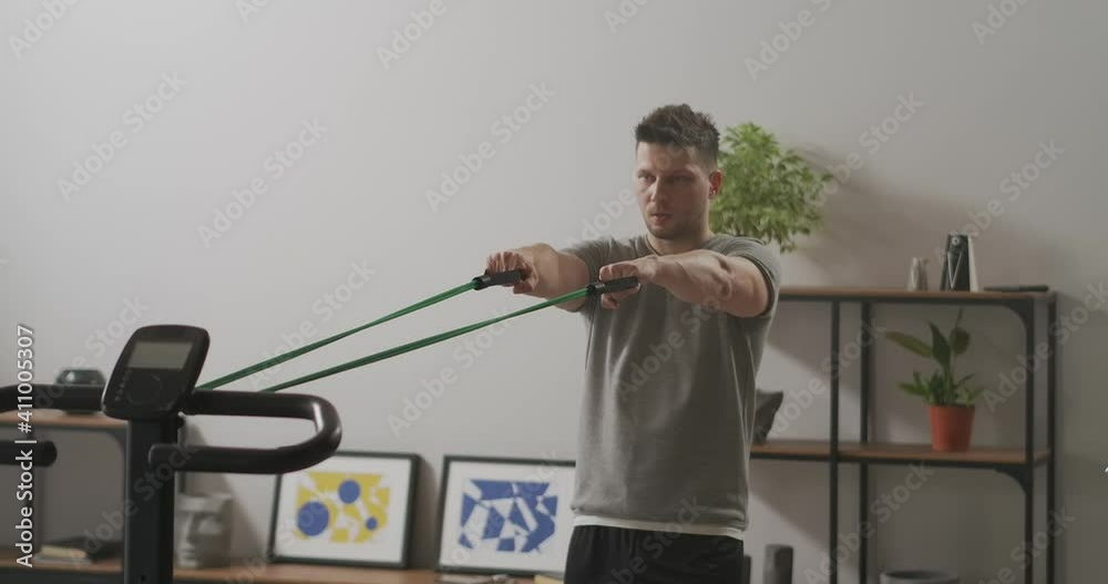 handsome man is doing exercises with sport rubber band in apartment, stretching tape and tensing muscles