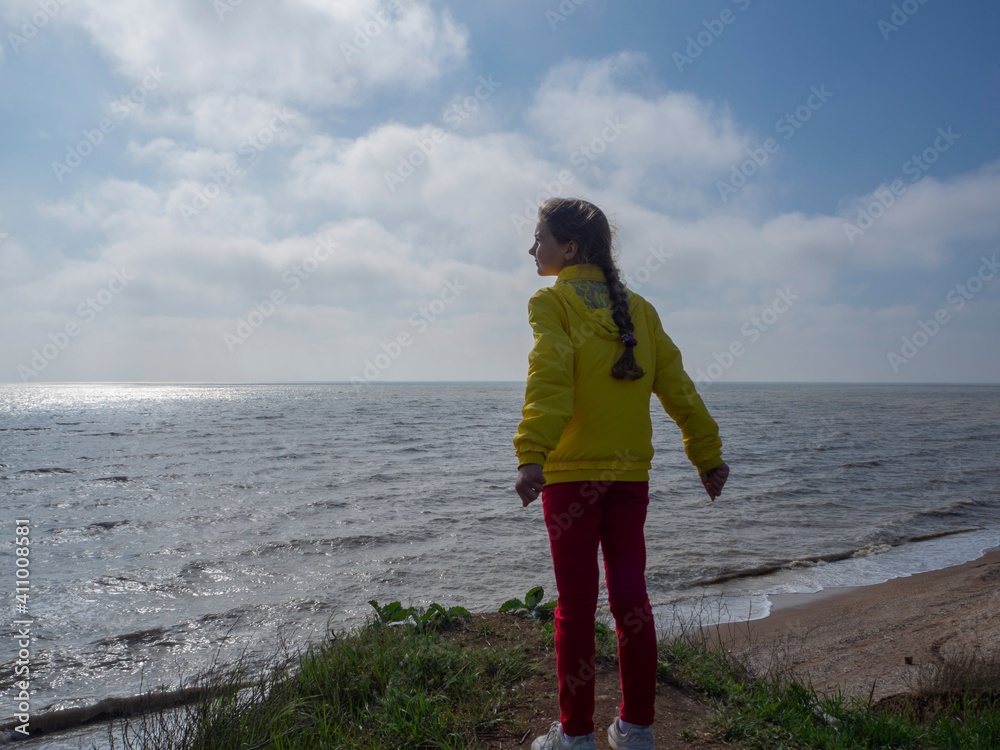A girl in a yellow windbreaker and red jeans stands on a high beach, looks into the distance, breathes clean air without a mask and enjoys a light breeze.