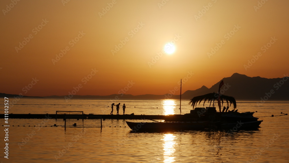 Orange sunset with boat near pier. Silhouettes of three people on a pier at sunset. Croatia, Makarska
