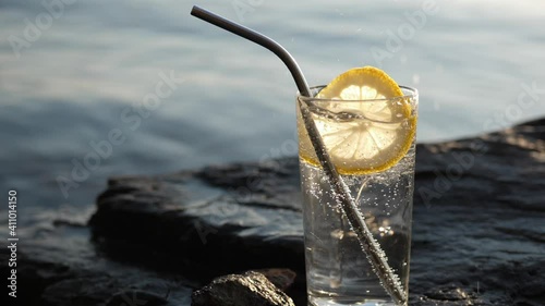 Slow motion male fingers toss round slice of lemon into glassful of cold clear soda water. Non-alcoholic drink in transparent tall glass stands on stony seashore under the bright sun. photo