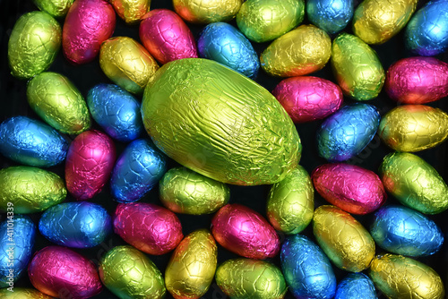 Pile or group of multi colored and different sizes of colourful foil wrapped chocolate easter eggs in pink, blue, yellow and lime green with a large green egg in the middle.