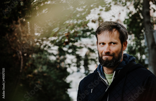 Handsome middle-aged Caucasian man portraited in the city, sitting on a bench with grey and black hair having a friendly look on his face, wearing a blue jacket and jeans. Fall colourful background.