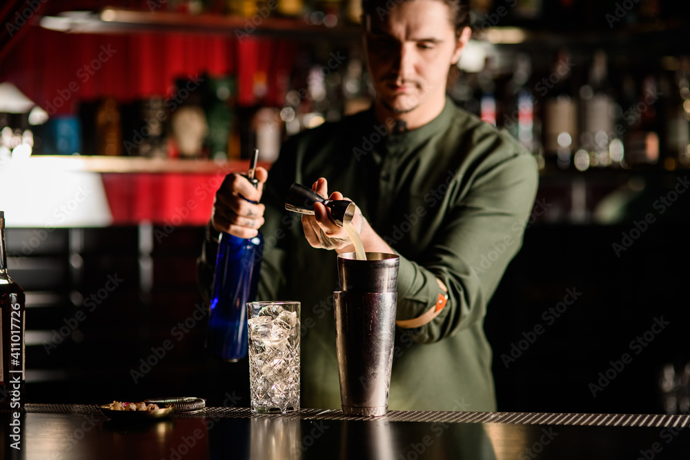 young male bartender gently pours jigger drink into steel glass of shaker.