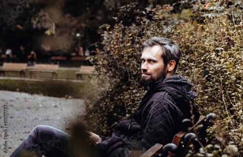 Handsome middle-aged Caucasian man portraited in the city, sitting on a bench with grey and black hair having a friendly look on his face, wearing a blue jacket and jeans. Fall colourful background.