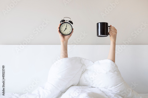 Woman showing arm raised up holding coffee cup and black alarm clock behind duvet in the bed room, Young girl with two hands sticking out from the blanket. wake up with fun in morning concept.