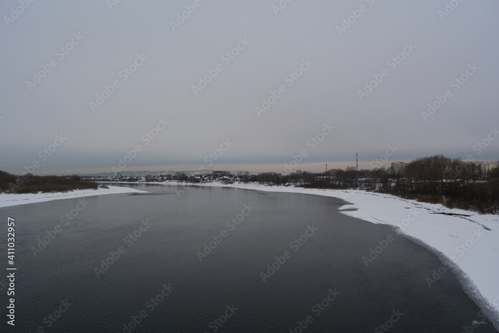 Winter landscape with unfrozen river and town on the bank in cloudy day