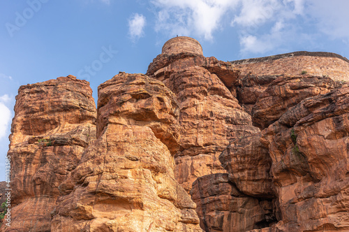 Badami, Karnataka, India - November 7, 2013: Cave temples above Agasthya Lake. Above reddish rock cliffs remnants of fortification with watch tower under blue cloudscape.