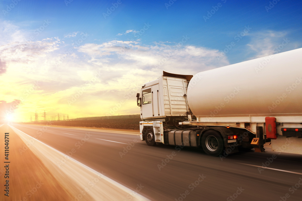 A fuel tanker truck shipping fuel on the countryside road in motion against a sky with a sunset