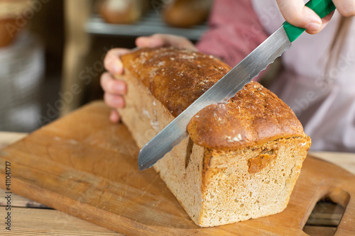 A new and young saleswoman in a bakery shop is slicing bread from a mold. For the first time in a new job, the girl starts cutting bread with a sharp knife with cloves.