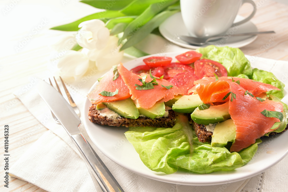 Whole meal sandwich with avocado and smoked salmon on lettuce and tomato salad, cutlery and coffee cup on a white painted wooden table, copy space