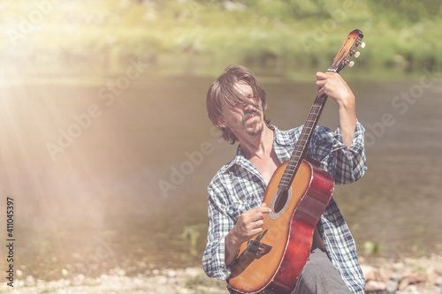  Rock musician man in an open shirt enthusiastically plays the old guitar in nature. Summer, clear day. Retro style, soft light. Musical concept. Portrait, close-up.