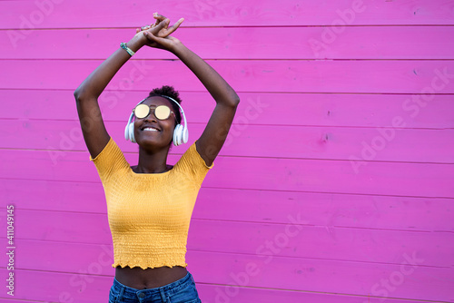 black young woman dancing on a fuchsia background, attractive afro girl with wireless headphone having fun with music in a party, copy space, vibrant colours, fashion addict concept photo