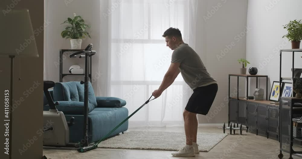 attractive man is doing physical exercise at home, training with rubber band, tensing arms muscles, keeping fit