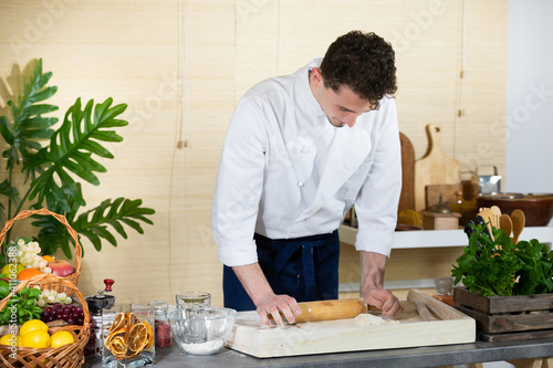 A young handsome chef is invited to the house as a helper, he rolls out a raw pizza dough on a dough lying on the kitchen counter. Professional cook at home.