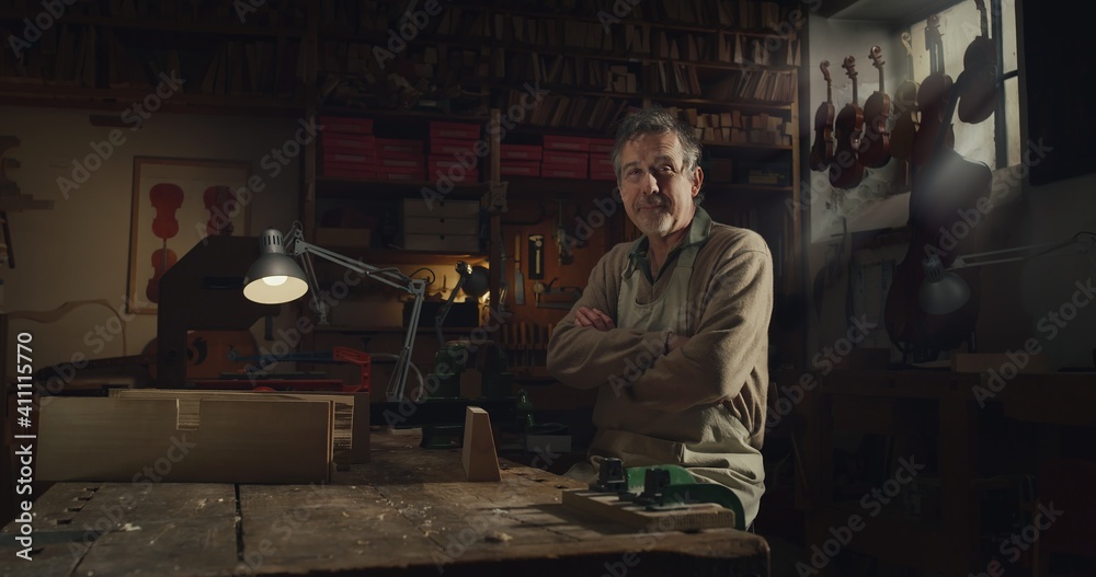 Cinematic shot of professional master artisan luthier is smiling satisfied with his job in camera while working on creation of handmade fine quality wood violin in creative workshop. Cinematic shot of professional master artisan luthier is smiling satisfied with his job in camera while working on creation of handmade fine quality wood violin in creative workshop.