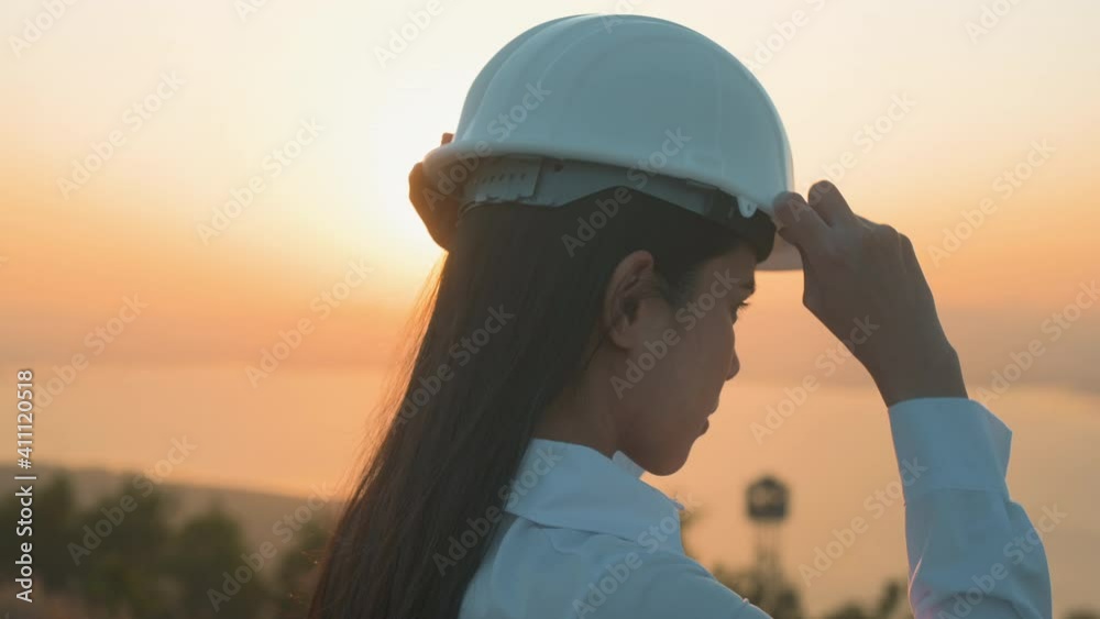 a woman engineer is putting a protective helmet on her head at sunset.