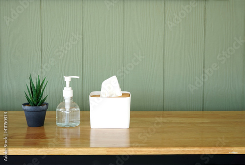 Tissue paper in a white box and hand alcohol gel bottle are on a wooden table. Showing hygiene and germs protection in house, restaurant, working space during pandemic. Grey panels in background.