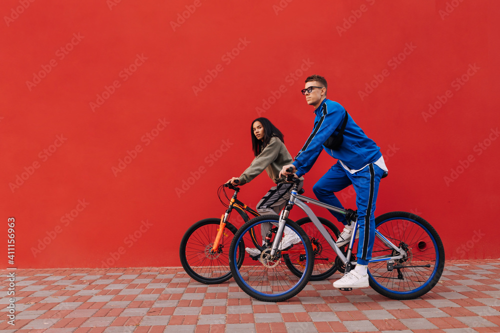 Naklejka premium Athletic couple of cyclists ride on a red background. Stylish young people walk on a bicycle on a background of a red wall on the city streets.