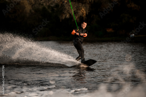 athletic man in wetsuit energetically rides on splashed river wave on wakeboard.