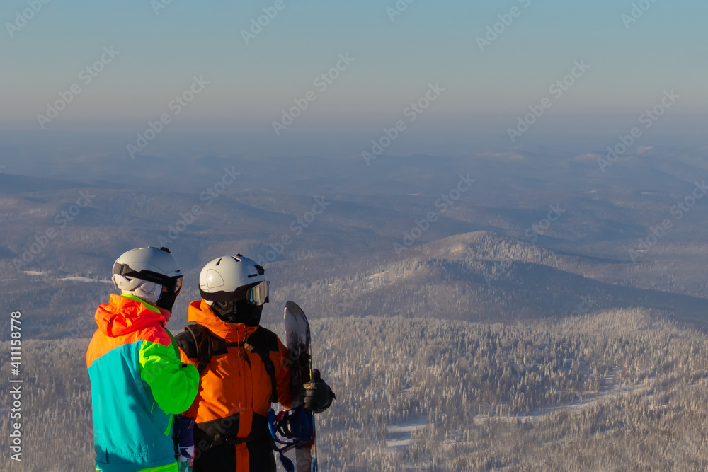 Two people in ski suits, masks and helmets stand on the top of a mountain on a sunny day. One of them is holding a snowboard