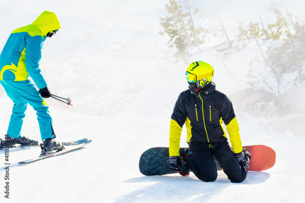 a skier talks to a snowboarder on a sunny winter day. A snowboarder sits on the snow, a skier stands